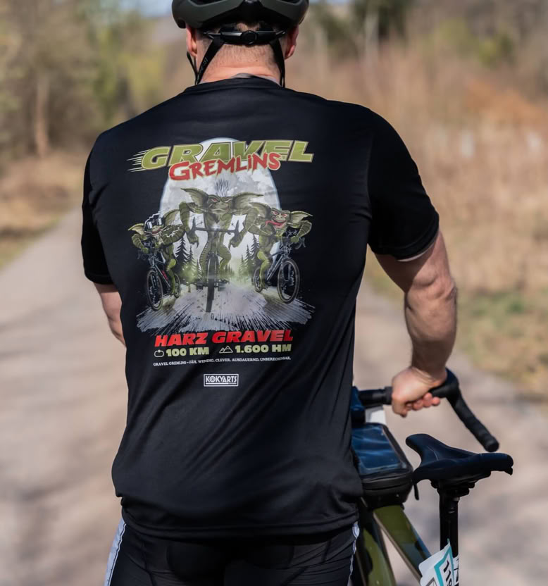 Eine Person mit schwarzem Helm und schwarzem T-Shirt steht auf einem Fahrradweg und hält ein Fahrrad. Im Hintergrund sind Bäume und ein blauer Himmel zu sehen.