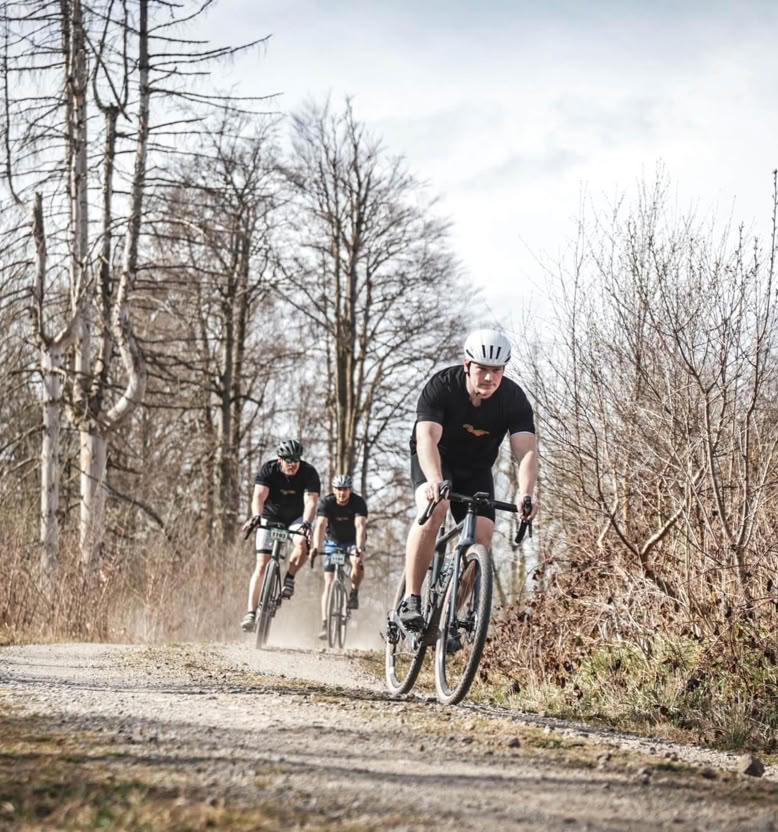 Drei Radfahrer fahren auf einem Waldweg, umgeben von kahlen Bäumen und Büschen.
