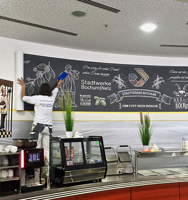 Person steht auf einer Leiter und arbeitet an einer Wandtafel in einem Café oder Restaurant. Auf der Theke stehen Pflanzen und Geräte.